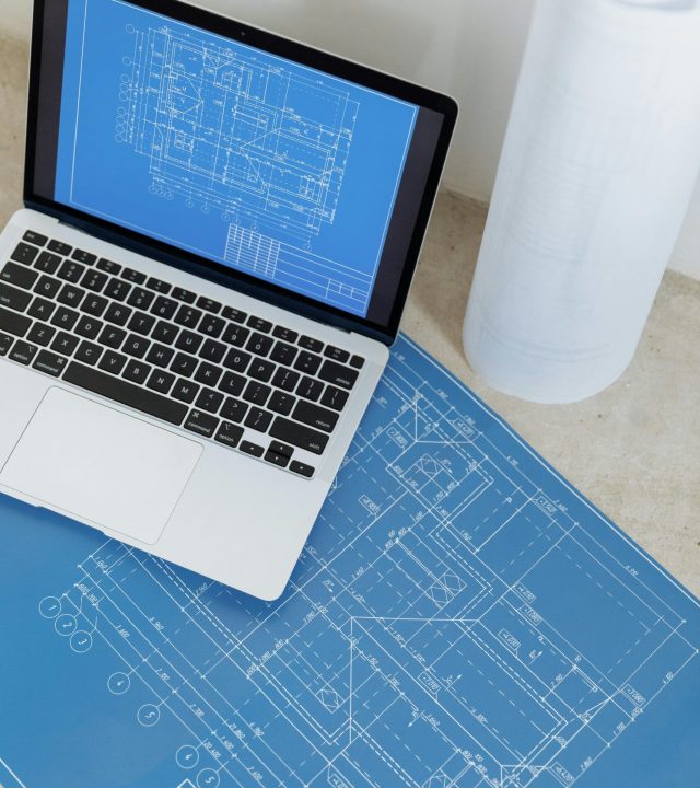 Laptop showing engineering blueprint with rolled paper and drawings on table.